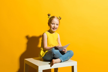 Wall Mural - Girl with red hair on a yellow background. Beautiful girl is sitting on the table and is holding a pencil and a white sheet in hands.