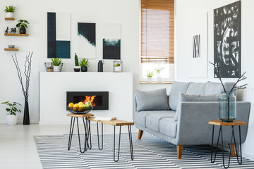 Real photo of a grey couch standing in front of a small wooden table in living room interior with a fireplace and posters on walls