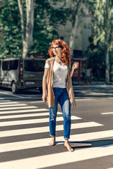 Adult happy pretty smiling caucasian redhead dressed in jeans barefoot female crossing city street on pedestrian or zebra crossing at sunny summer day.