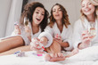 Blurry image of caucasian three women 20s wearing bathrobe sitting in big bed in posh apartment or hotel room, and painting nails during hen party