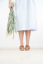 A Woman In A Light Blue Striped Skirt Holding A Bouquet Of Dried Grass Standing In A  Neutral Setting Wearing Tanned Brown Leather Clogs.