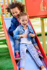 two excited screaming multiethnic little kids sliding down from hill at playground