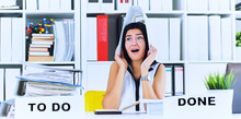 Funny Overworked Girl With A Folder On Her Head Sitting In The Workplace Cluttered With Folders. Reaction Of A Subordinate When The Boss Orders Her To Do Too Much.