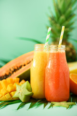Wall Mural - Juicy papaya and pineapple, mango, orange fruit smoothie in two jars on turquoise background. Detox, summer diet food, vegan concept. Copy space. Fresh juice in glass bottles over green palm leaves.