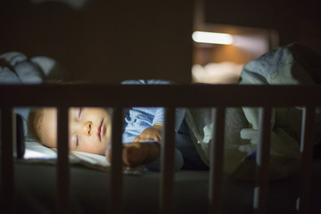 Wall Mural - Adorable newborn baby boy, sleeping in crib at night. Little boy in white pajamas taking a nap in dark room. Bedroom interi?r with lamp and teddy bear .