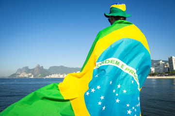 Wall Mural - Brazilian football fan wrapped in giant flag and hat in front of Ipanema Beach city skyline Rio de Janeiro.