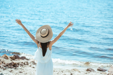 Wall Mural - rear view of woman in straw hat and white dress standing with wide arms by sea