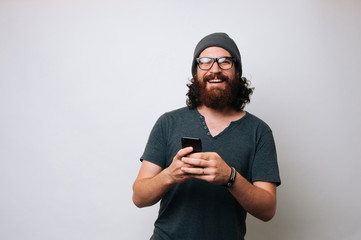 Portrait of a happy young bearded hipster man holding mobile phone while standing and looking at camera over white background