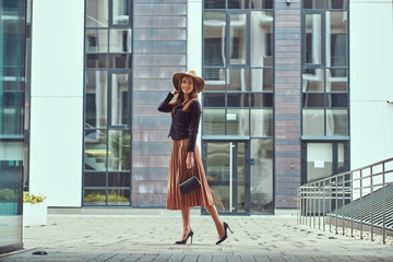 Happy fashion elegant woman wearing a black jacket, brown hat and skirt with a handbag clutch walking on the European city center.