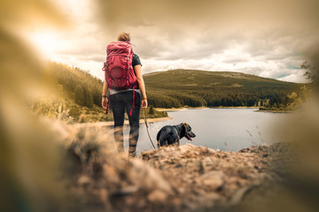 young woman with backpack and german shepherd dog puppy standing on mountain in front of forest and lake