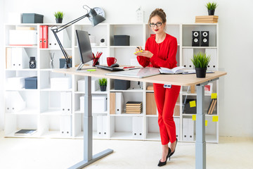 A young girl is standing near a table in the office, holding stickers and a black marker. The girl works with a computer, notepad and a magnetic board.