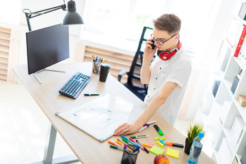 A young man in glasses stands near a computer desk and is talking on the phone. Before him lies a magnetic board and markers. On the neck, the guy's headphones hang.
