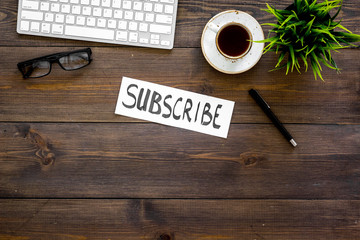 Email subscribe concept. Hand lettering subcribe on work desk with plant, glasses, computer keyboard, cup of coffee on dark wooden background top view copy space