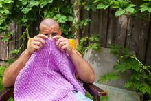 A Young Bald Man Sits On An Armchair And Knits A Sweater From Natural Lilac Wool Threads At The Dacha, In The Background A Wooden Fence, Grape Bushes