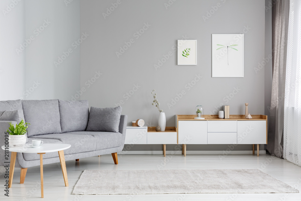 Real photo of a spacious living room interior with gray sofa standing between a wooden table and white cupboards - obrazy, fototapety, plakaty 
