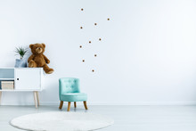 Small Light Blue Armchair For Kid Standing In White Room Interior With Stars On The Wall, White Rug And Cupboard With Books, Teddy Bear And Fresh Plant. Empty Space For Your Crib