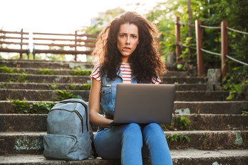 Canvas Print - Photo of upset confused woman sitting on stairs in park on summer day, and using silver laptop with disappointment