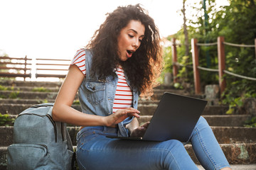 Canvas Print - Photo of excited brunette woman sitting on stairs in park on summer day, and using silver laptop with surprise