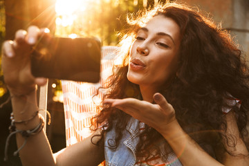 Wall Mural - Photo of seductive flirty woman 18-20 smiling and blowing air kiss at cell phone, while taking selfie photo during rest in lounge chair in park on sunny day