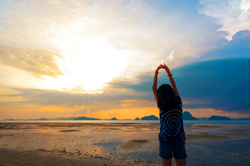 Wall Mural - Silhouette of young woman stretching arms standing on tropical beach with sunset or sunrise sky background. Joyful Asian girl holding hands and breathing fresh air outdoors in the morning, copy space.