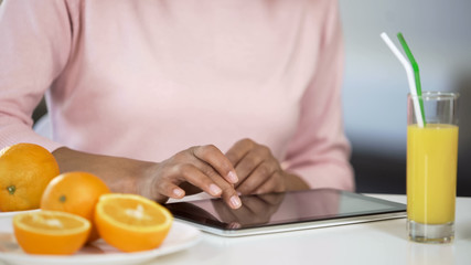 Girl typing on tablet, oranges and fresh juice on table, blogging about dieting