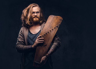 Wall Mural - Redhead hipster male with long luxuriant hair and full beard dressed in casual clothes holds a Russian traditional musical instrument - gusli. Isolated on the dark background.