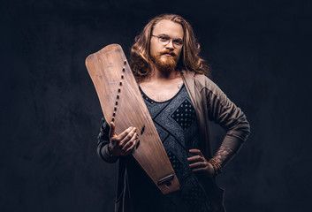 Wall Mural - Redhead hipster male with long luxuriant hair and full beard dressed in casual clothes holds a Russian traditional musical instrument - gusli. Isolated on the dark background.