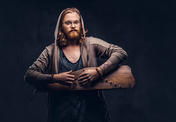 Wall Mural - Redhead hipster male with long luxuriant hair and full beard dressed in a hoodie and t-shirt playing on a Russian traditional musical instrument - gusli. Isolated on a dark background.