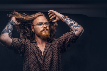 Wall Mural - Tattoed redhead man hipster corrects his lush hair dressed in a brown shirt, standing at a studio. Isolated on a dark background.