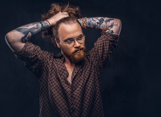 Wall Mural - Tattoed redhead man hipster corrects his lush hair dressed in a brown shirt, standing at a studio. Isolated on a dark background.