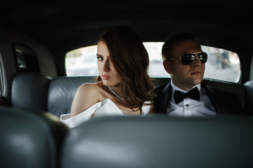 Stunning bride and groom in black sunglasses sit inside a retro car