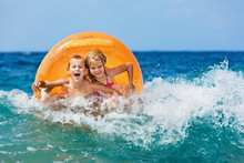 Happy Kids Have Fun In Sea Surf On Beach. Joyful Couple Of Children On Inflatable Ring Ride On Breaking Wave. Travel Lifestyle, Swimming Activities In Family Summer Camp. Vacations On Tropical Island 