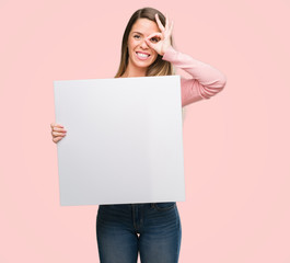 Sticker - Beautiful young woman holding advertising banner with happy face smiling doing ok sign with hand on eye looking through fingers