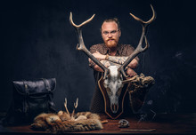 Redhead Taxidermist Hipster Male In Sunglasses Dressed In A Brown Shirt, Standing Near A Table With Handmade Trophy, Owl Scarecrow, And The Fox Skin. Isolated On The Dark Background.