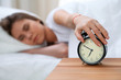 © rogerphoto - Sleepy young woman stretching hand to ringing alarm willing turn it off. Early wake up, not getting enough sleep concept