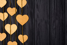 Flat Lay Border Of Golden Hearts And Beads. Festive Decor On A Black Wooden Background. View From Above.