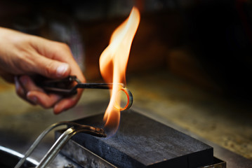 Wall Mural - goldsmith hands glow a golden ring in a gas flame, close up with copy space in the dark background