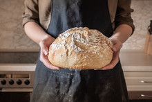 Hands Holding Big Loaf Of White Bread. Female In Black Apron In Home Kitchen Background With Wheat Bread