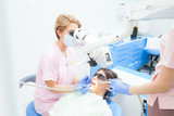 Fototapeta Panele - Young female dentist treating root canals using microscope at the dental clinic. Young woman patient lying on dentist chair with open mouth. Dentist wearing mask and gloves