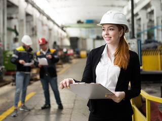 A young successful woman engineer working at a modern plant. In the background 2 workers. The girl in a white helmet holding technical documentation.