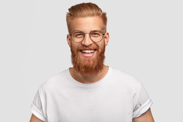 People, youth, positiveness concept. Happy male with long thick ginger beard, has friendly smile, rejoices having day off for his hobby, expresses happiness, stands alone against white wall.