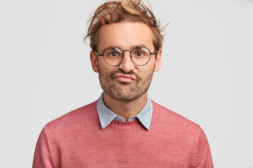 Wall Mural - Puzzled displeased male curves lips, looks doubtfully into camera, feels hesitation, dressed in pink sweater, has curly hair, poses against white background. Discontent Caucasian man indoor.