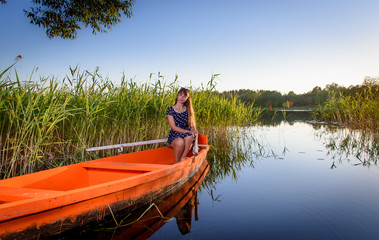  Young simpotic girl in a summer blue dress is sitting in a boat on the shore of a beautiful lake.