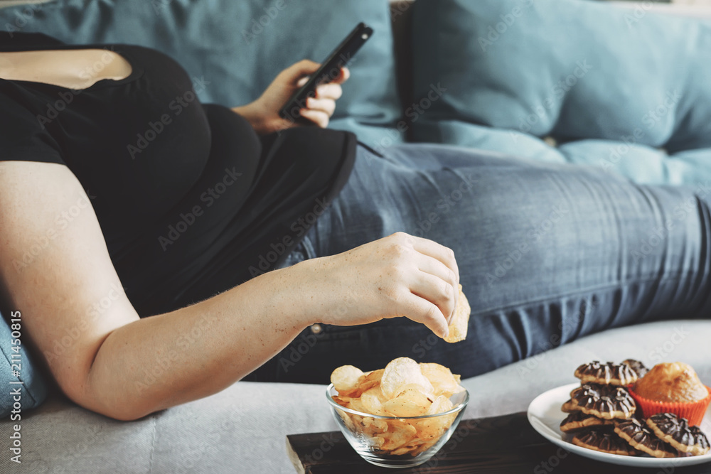 g, sedentary lifestyle, compulsive overeating. Obese woman laying on sofa with smartphone eating chips - obrazy, fototapety, plakaty 