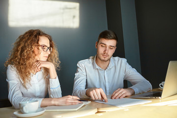 Candid shot of attractive young male employee and his female colleague with long wavy hair sitting at desk with papers, having discussion, brainstorming, sharing ideas with each other in office