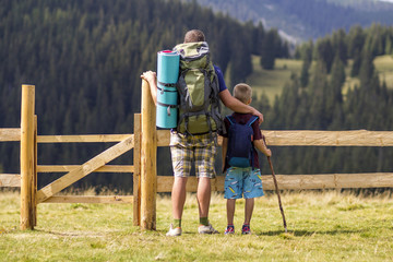 Wall Mural - Dad and son child with tourist backpacks at low wooden fence on green mountains covered with pine forest background. Active lifestyle, tourism tradition, family relations and weekend activity concept.