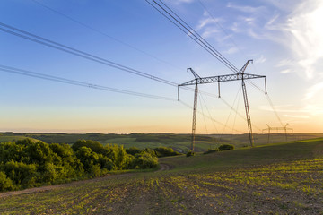 Beautiful wide panorama of high voltage lines and power pylons stretching through spring fields above group of green trees at dawn or sunset. Transmission and distribution of electricity concept.