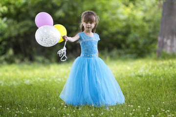 Beautiful little blond long-haired girl in nice long blue evening dress looking like princess looks in camera holding colorful balloons standing in blooming field on blurred green trees background.