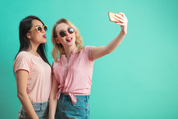 Two diverse teenage girls in summer shorts and sun glasses making selfie, bulging out their lips on blue background with copyspace.