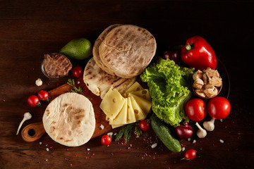 Plain pita on a cutting board with fresh vegetables and cheese on a wooden background, flat lay, selective focus.